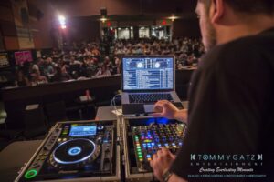 A DJ stands at a console with turntables and a laptop, mixing music for an engaged audience in a dimly lit venue.
