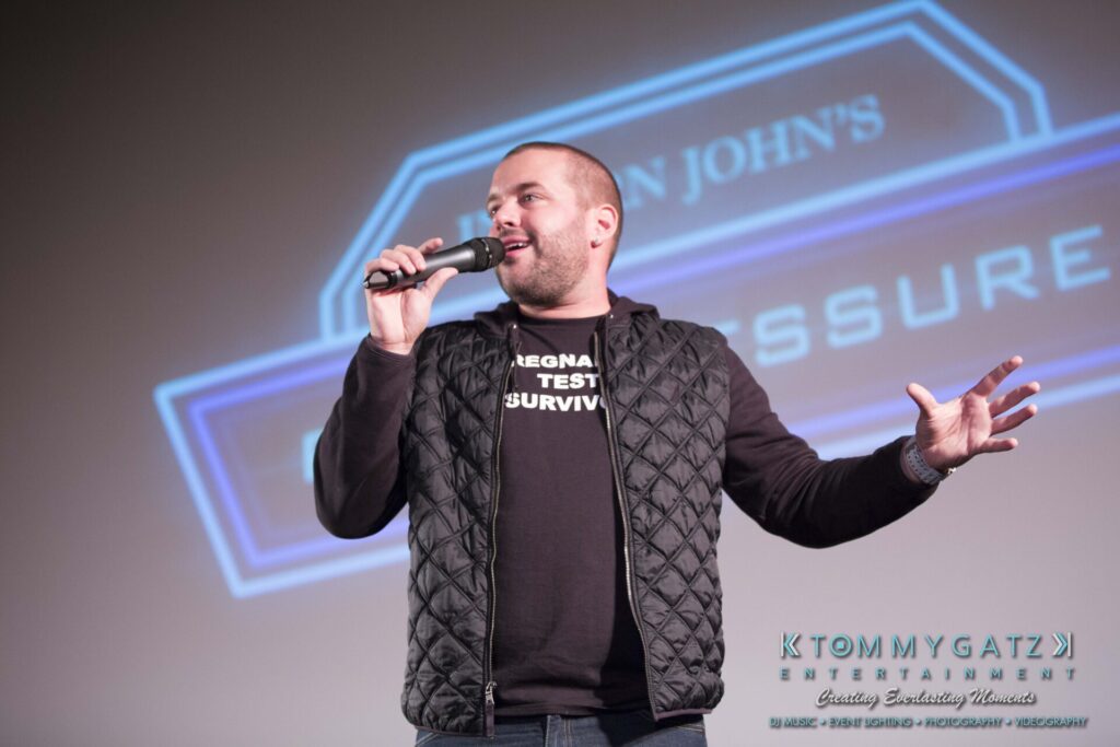 A person in a black quilted jacket speaks on stage with a microphone. A large logo is projected in the background.