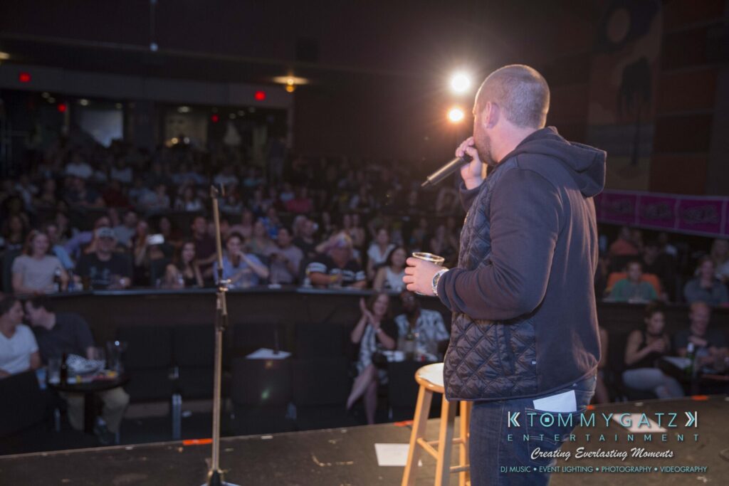 A person with a microphone addresses a seated audience in a dimly lit venue. A spotlight shines from the stage, and there are visible logos at the bottom of the image.