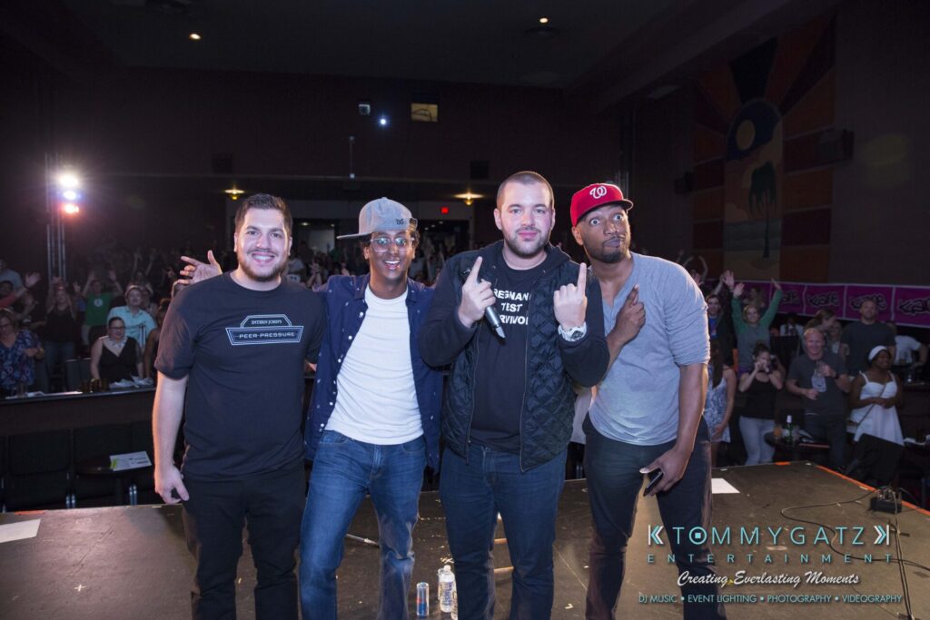 Four men standing on stage in a theater setting, with an audience in the background. Lighting and photography are credited to Tommy Gatz Entertainment.
