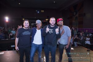 Four men standing on stage in a theater setting, with an audience in the background. Lighting and photography are credited to Tommy Gatz Entertainment.