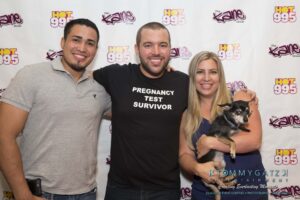 Three people standing together smiling in front of a backdrop with logos, one person is holding a small dog.