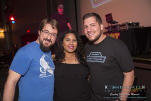 Three people smiling in a dimly lit indoor event space, standing in front of a screen and DJ equipment.