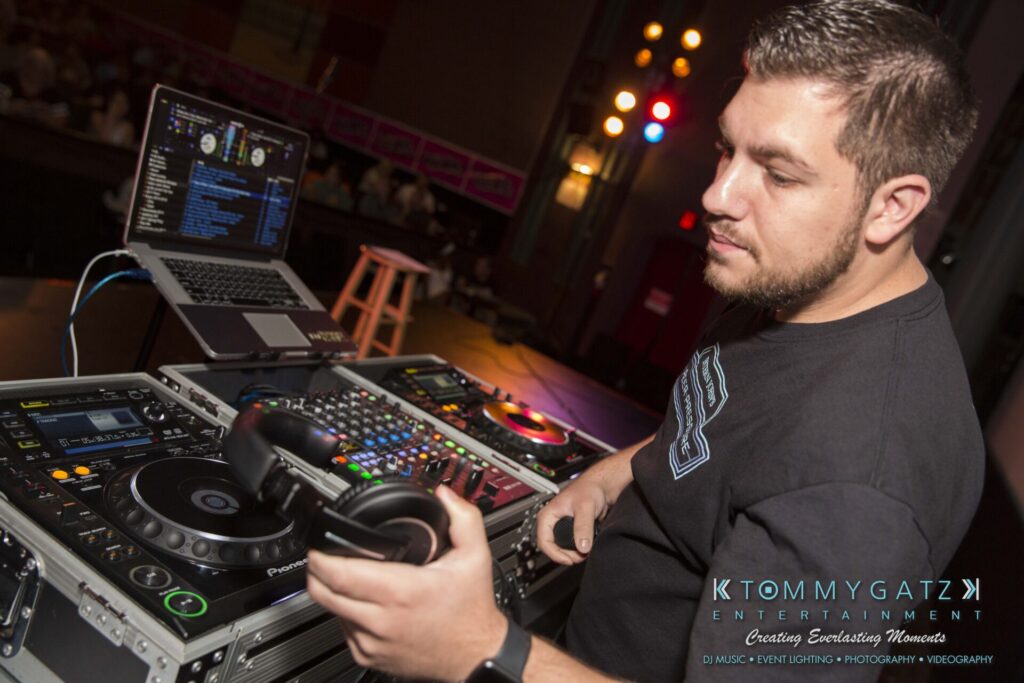 DJ at a mixing console adjusting equipment, with a laptop displaying music software in the background.