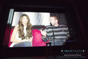 A presentation screen shows two people sitting on a couch, with one speaking into a microphone. A person operates equipment in the foreground.