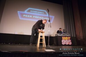 A man is standing and writing on a notepad on a stool on stage, with a microphone next to him and a background screen displaying "Intern John's Peer Pressure.
