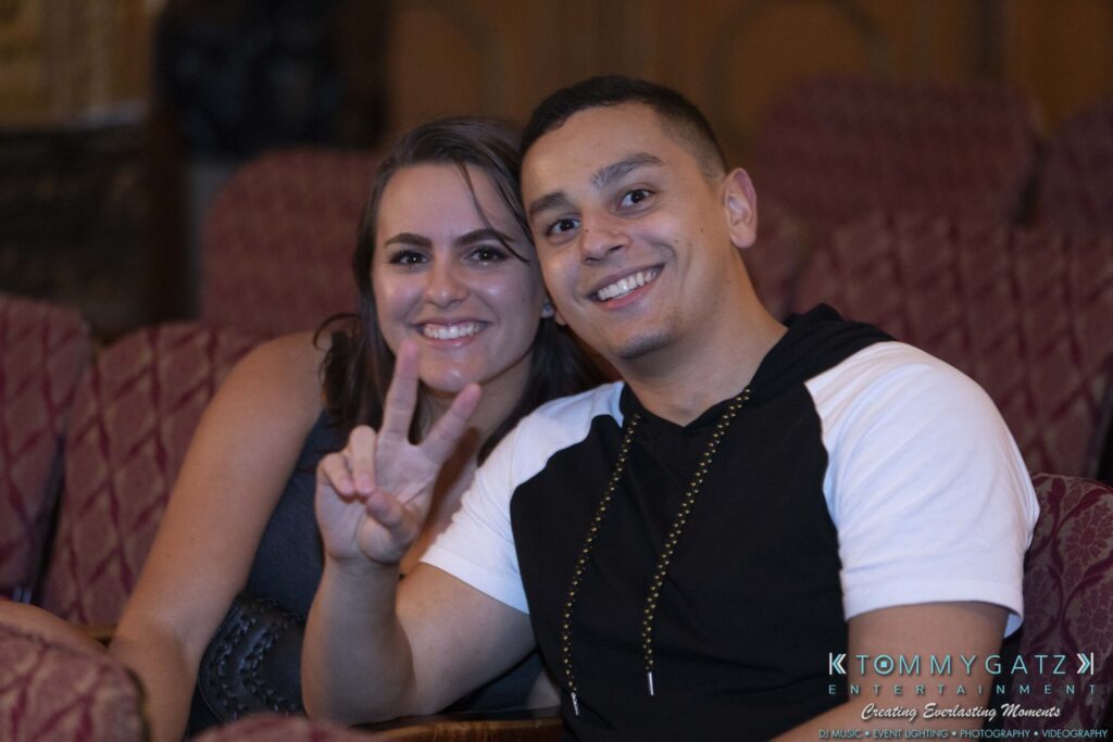 A person with a peace sign and another smiling, sitting on red patterned chairs in a dimly lit room.