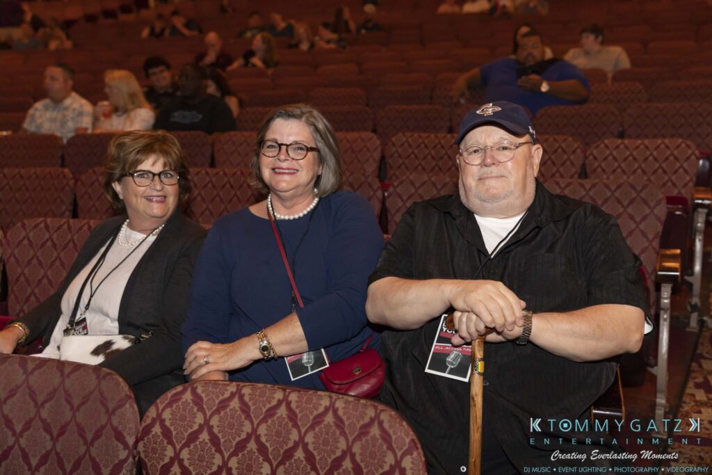 Three people sitting in a theater, smiling at the camera. Two women are on the left, and a man with a cane is on the right. Seats and more people in the background.
