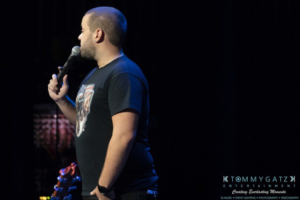 A man in a black t-shirt holds a microphone on stage, performing stand-up comedy, with a guitar visible in the background.