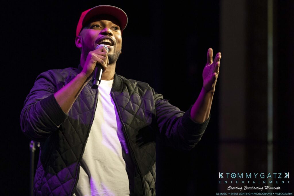 A person wearing a quilted jacket and red cap holds a microphone and gestures with the other hand on stage.