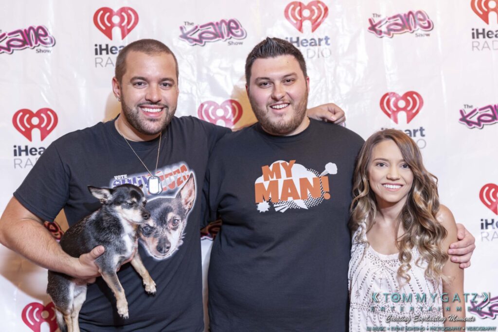 Three people pose for a photo on a red carpet backdrop with iHeartRadio logos. One man holds a small dog.