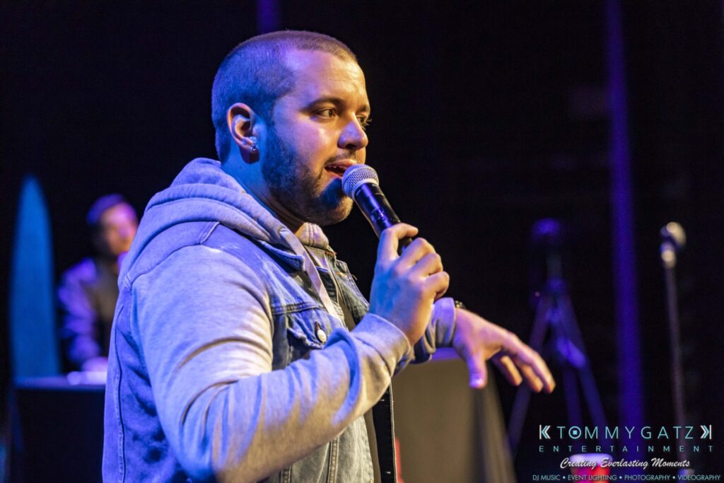 A man in a denim jacket speaks into a microphone on stage, with another person blurred in the background.