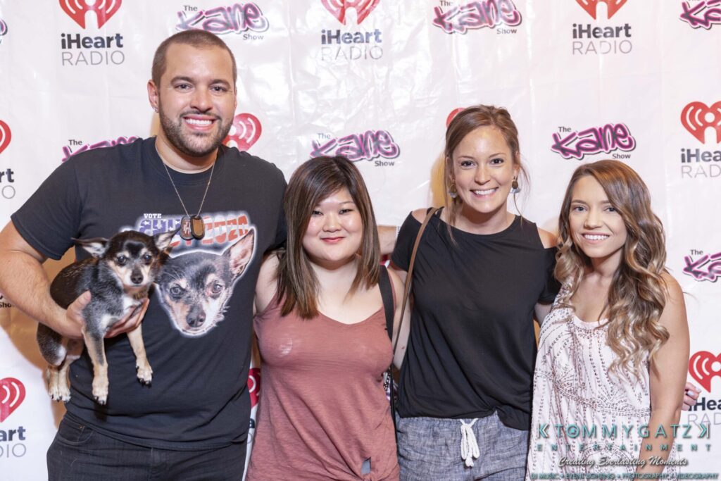 Four people stand in front of an iHeart Radio backdrop, with one person holding a small dog.