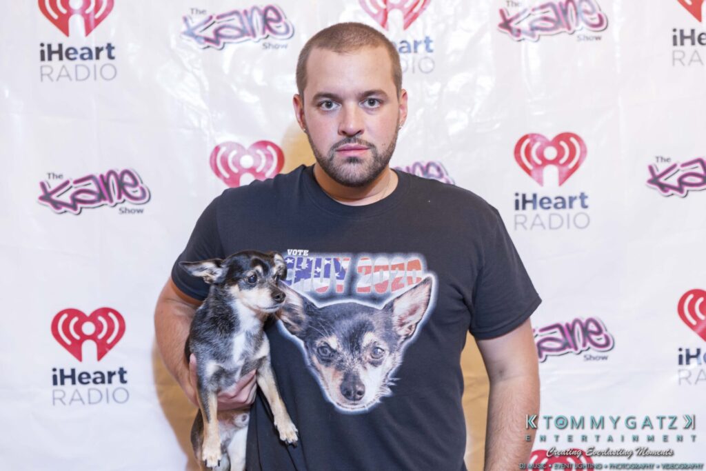 A man stands in front of a backdrop holding a small dog. He's wearing a T-shirt with a similar dog image. The backdrop features iHeartRadio and "The Kane Show" logos.