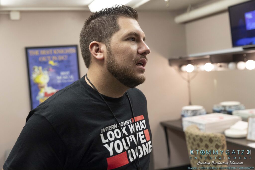 A man with a beard wearing a black T-shirt with a motivational message stands in a room with makeup mirrors and posters on the wall.