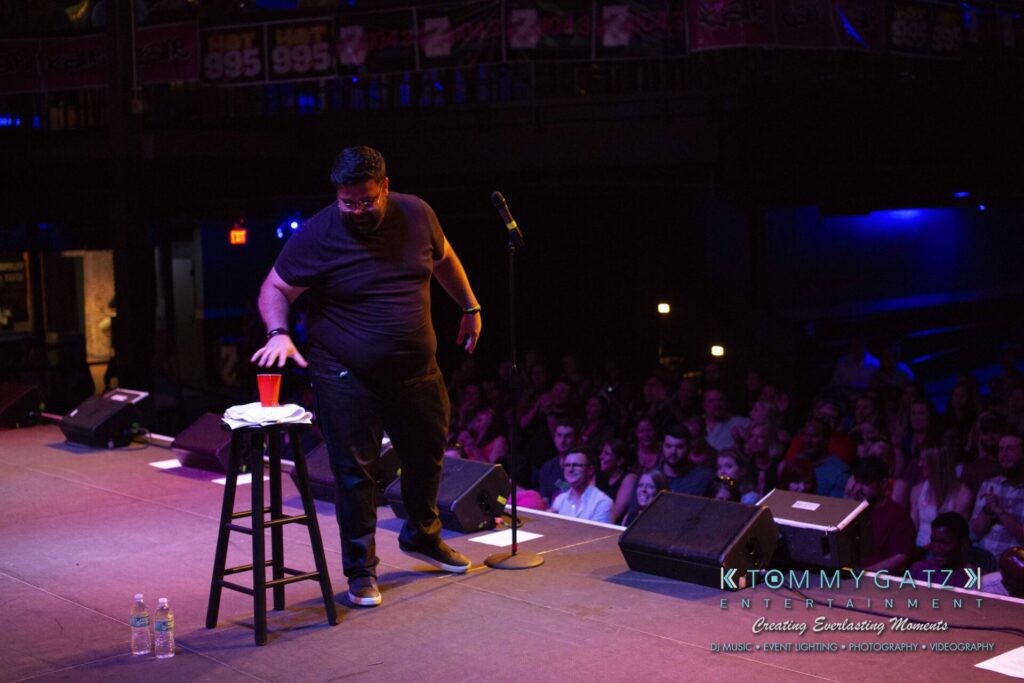 A performer on stage reaches for a cup on a stool, with an audience seated in the background.
