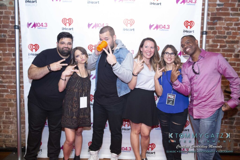 A group of six people posing in front of a iHeartRadio and Z104.3 backdrop, making peace signs and smiling.