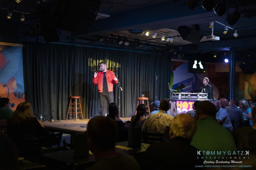 A comedian performs on stage with a microphone in hand. A DJ stands nearby at a booth. The audience watches from tables in a dimly lit comedy club.