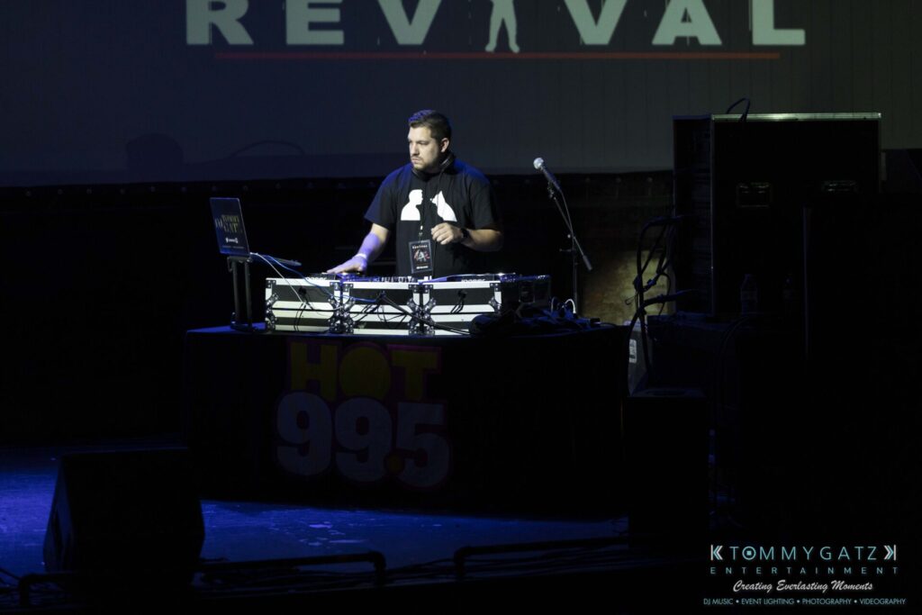 A DJ stands behind a mixer and laptop on a dimly lit stage with "REVIVAL" partially visible in the background.