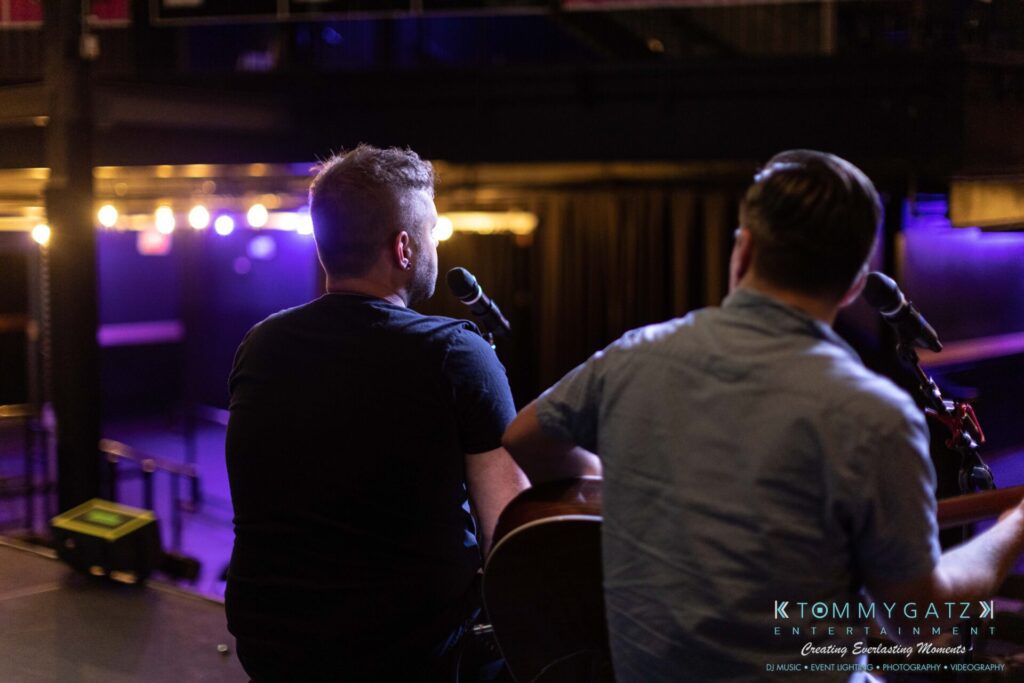 Two musicians on stage with microphones, viewed from behind. Stage lights illuminate the background.