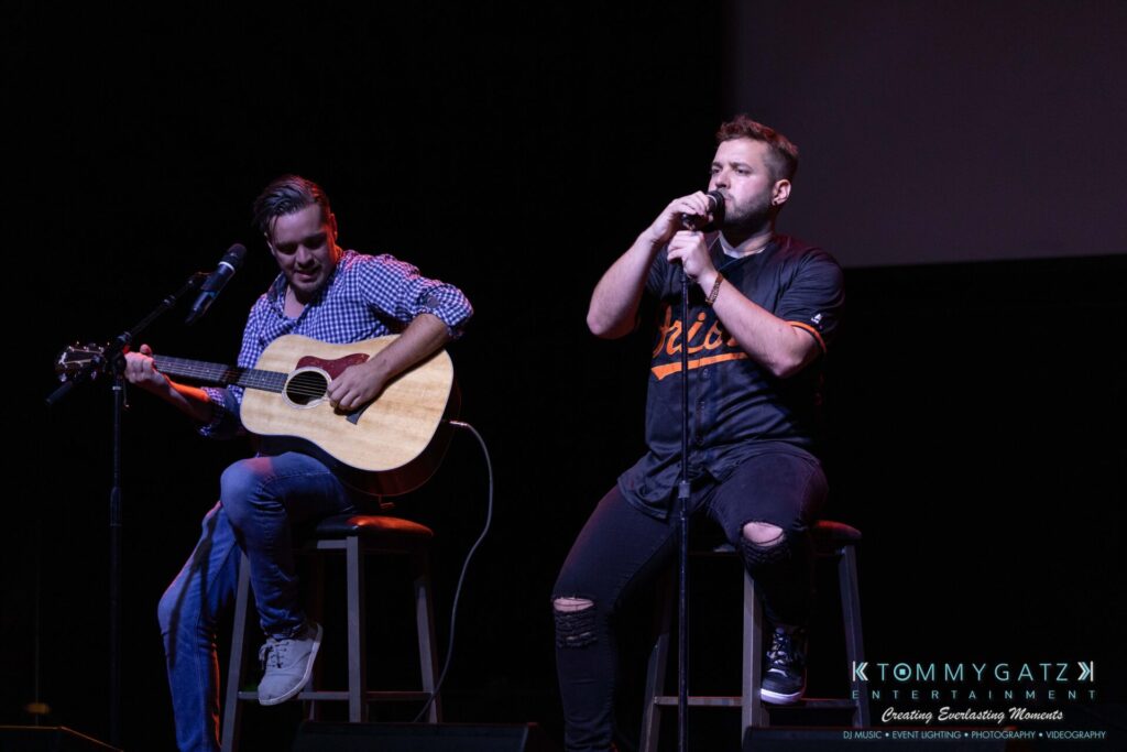 Two musicians perform on stage; one plays an acoustic guitar and the other sings into a microphone. Both are seated on stools, against a dark backdrop.