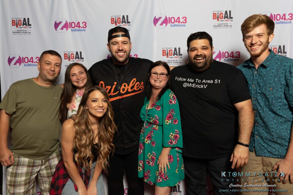 A group of seven people smiling and posing together in front of a backdrop with radio station logos.