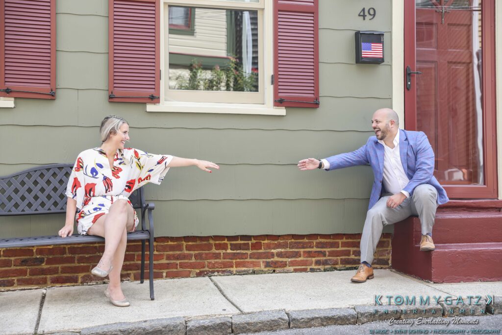 A woman and a man sit on a bench and a ledge, reaching out towards each other in front of a green, brick-trimmed building with a small American flag.