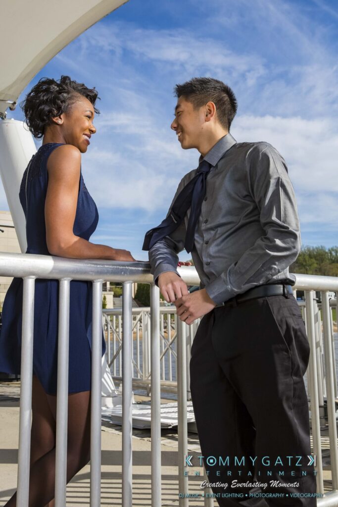 A man and woman smile at each other while leaning on a railing outdoors on a sunny day.