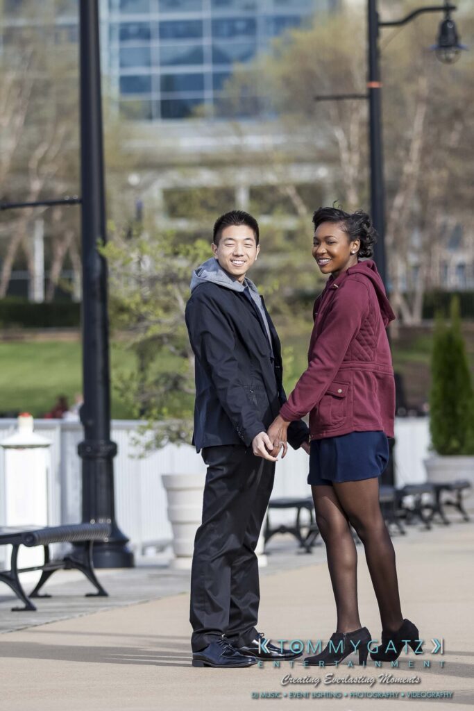 A couple stands outdoors holding hands, smiling at the camera. Urban setting with benches and trees in the background.