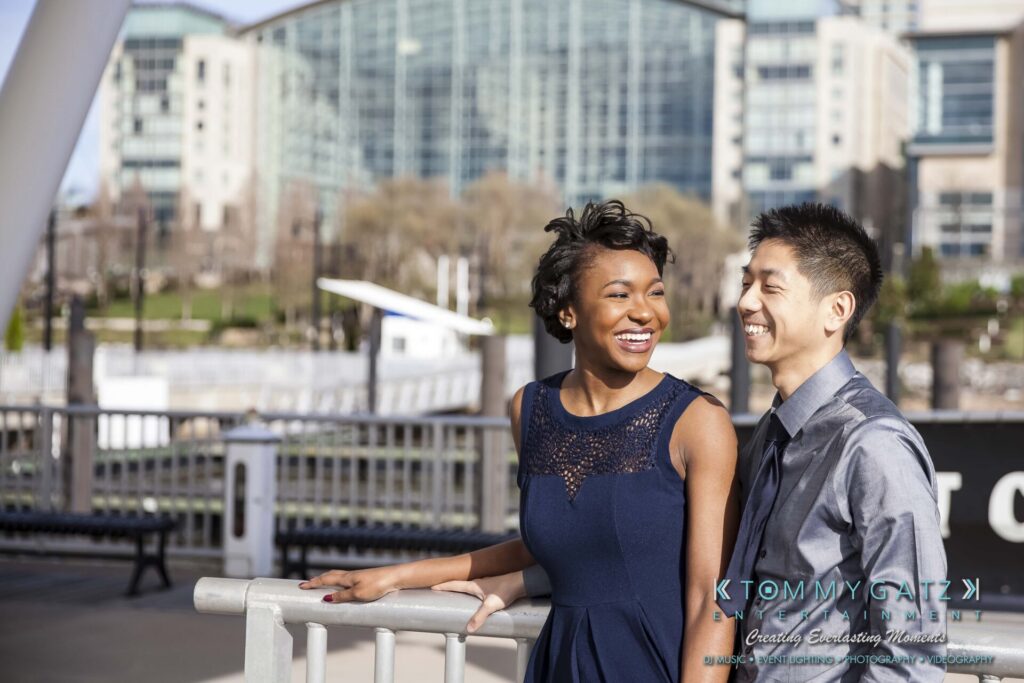 A woman and a man stand side by side smiling outdoors near a railing, with a cityscape visible in the background.