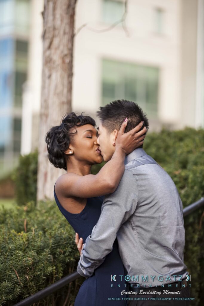 A couple kisses outdoors, with greenery and a building in the background.