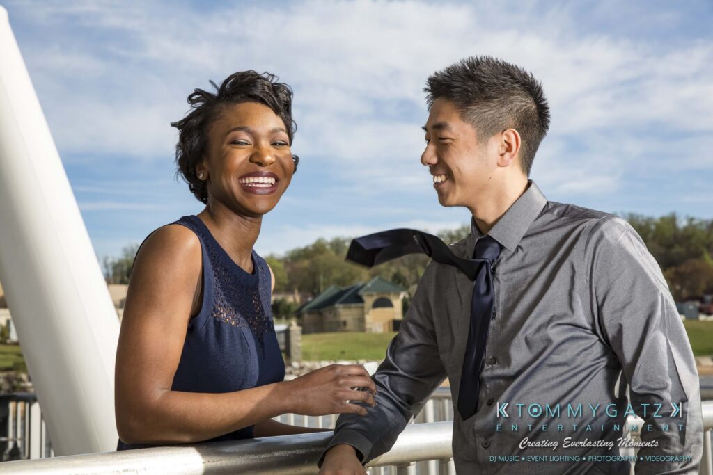 A man and woman smiling, standing outdoors by a railing. The man is in a tie, the woman in a sleeveless dress.