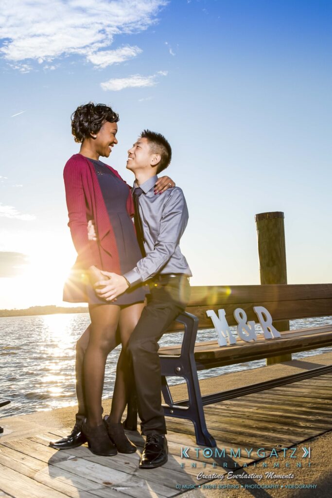 A couple sits on a bench at a pier by the water, smiling at each other as the sun sets.