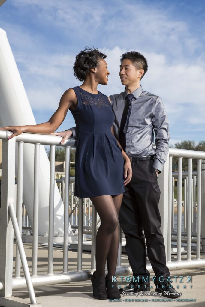 A couple stands together on a bridge. The woman leans on a railing wearing a dark blue dress, while the man stands beside her wearing a gray shirt and tie. Both gaze at each other.