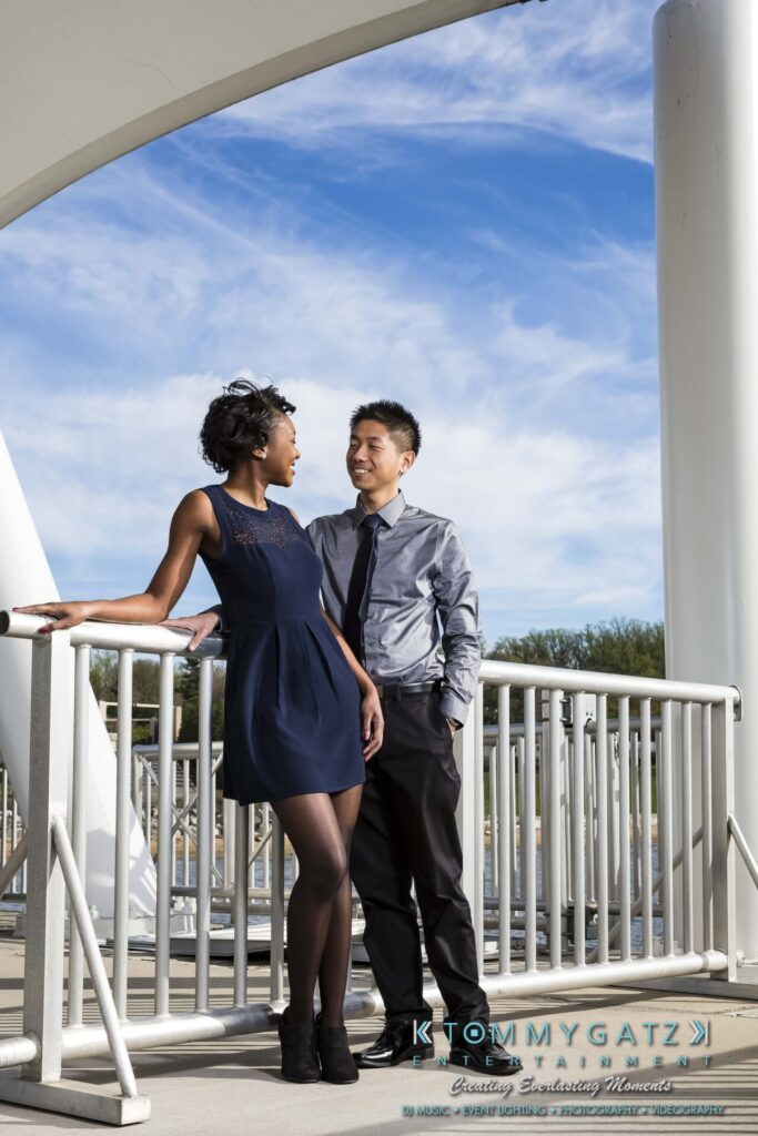 A man in a shirt and tie stands next to a woman in a blue dress, leaning against metal railings under a white arch. The sky is clear and blue.