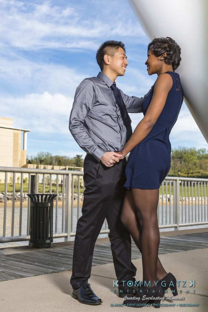 A couple stands on a pier, smiling and holding hands. The background features water and buildings.