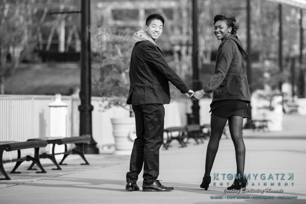 A man and a woman smile while holding hands and walking on a paved path lined with benches and lampposts.