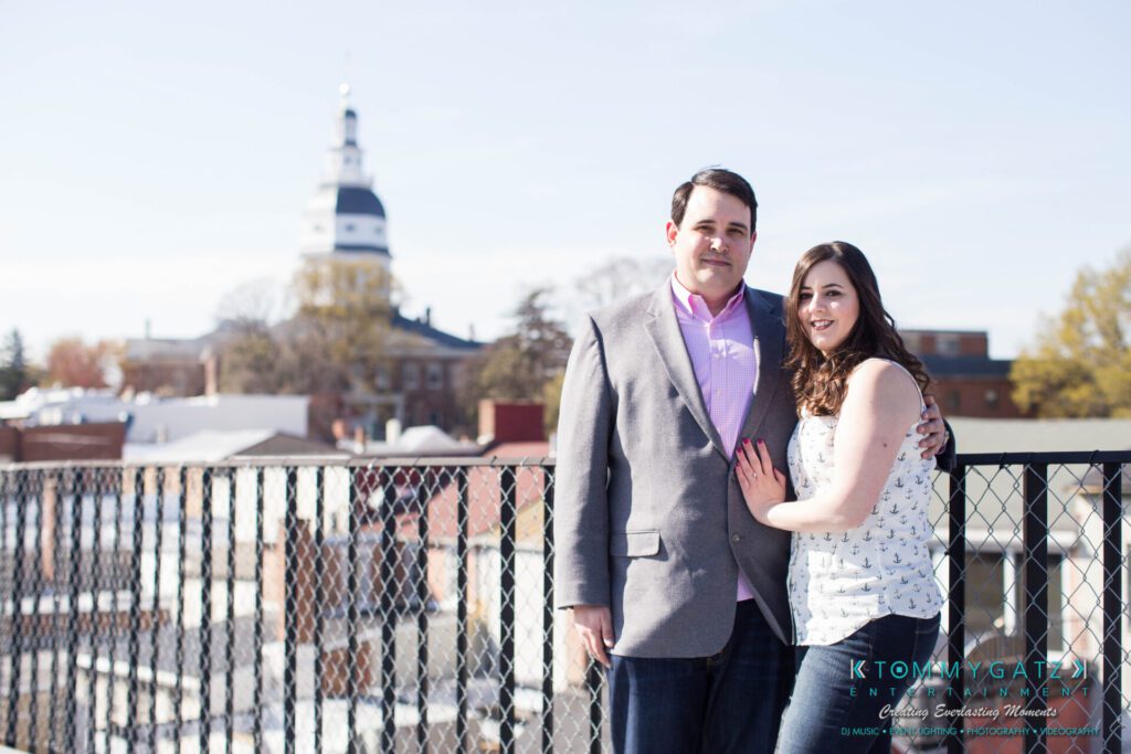 A couple stands together on an outdoor balcony overlooking a cityscape with a domed building in the background.