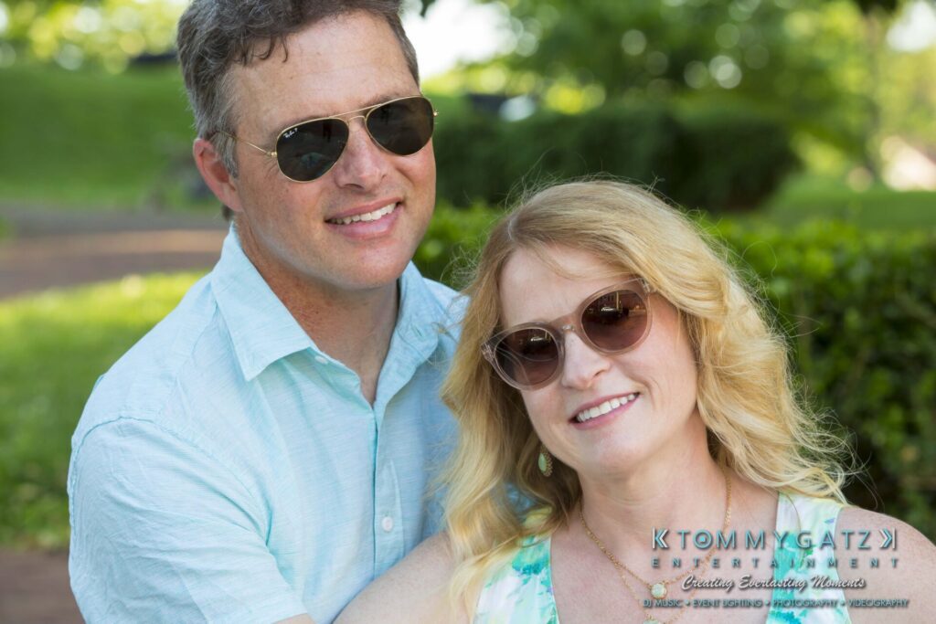A man and woman wearing sunglasses are smiling outdoors, with greenery in the background. The man is wearing a light blue shirt, and the woman is wearing a floral dress.