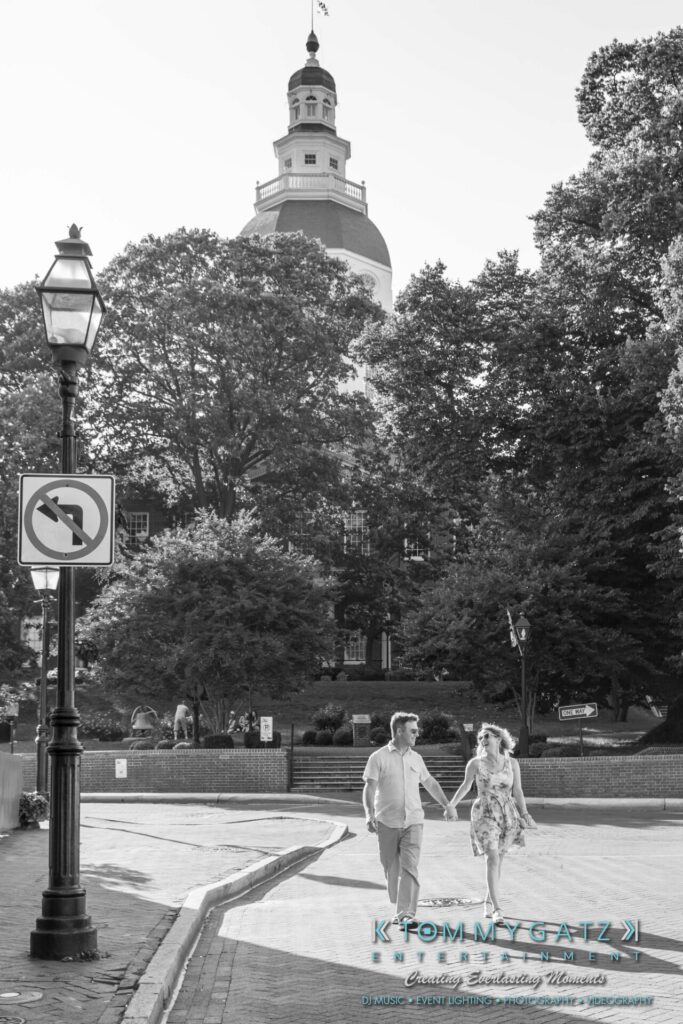 A couple holding hands walks across a street near a large, domed building surrounded by trees.