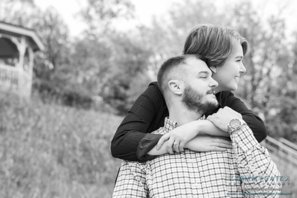 A woman embraces a man from behind, both smiling. They are outdoors in a grassy area with trees, near a gazebo. Black and white photo.