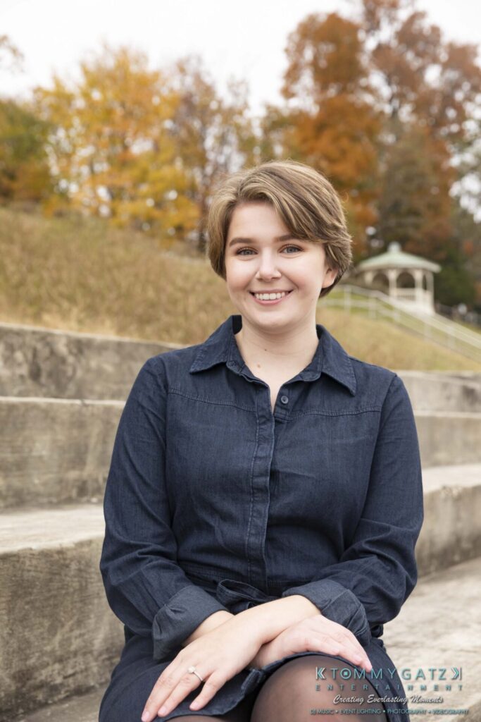 A smiling person with short hair sits on outdoor steps wearing a dark dress. Autumn trees and a gazebo are in the background.