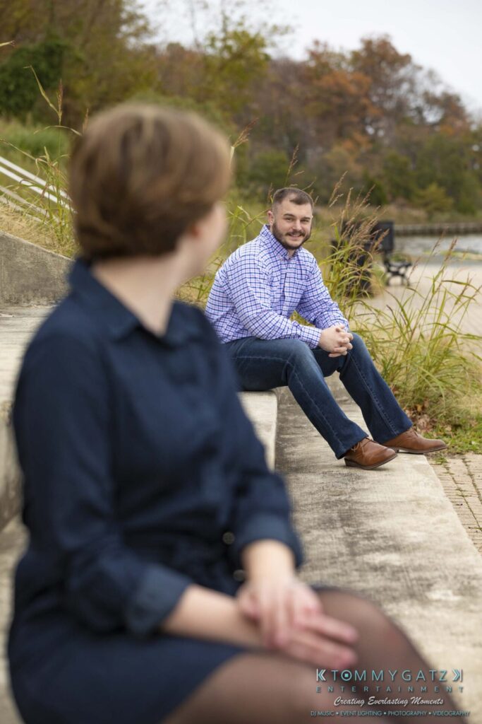 A person in a blue dress is sitting on steps in the foreground, facing away. In the background, a person in a checked shirt is sitting on steps, facing forward. Grass and trees are nearby.