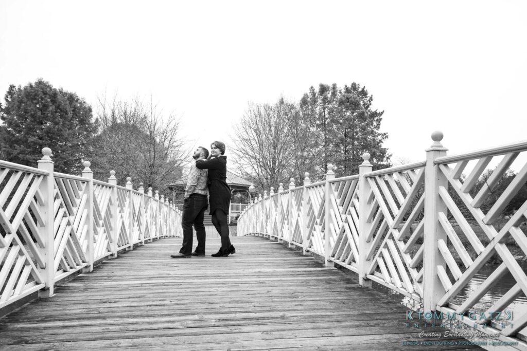 Two people standing on a wooden bridge, embracing with trees in the background on a cloudy day.