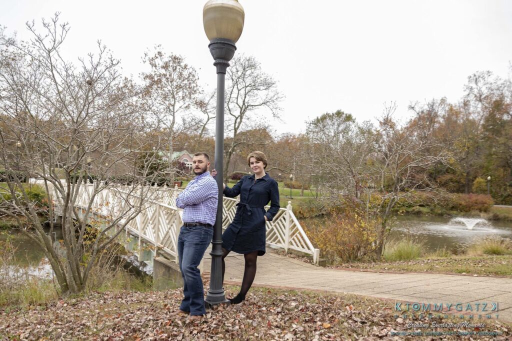 Two people stand back-to-back leaning against a lamppost on a bridge. Trees and a pond with a fountain are in the background.