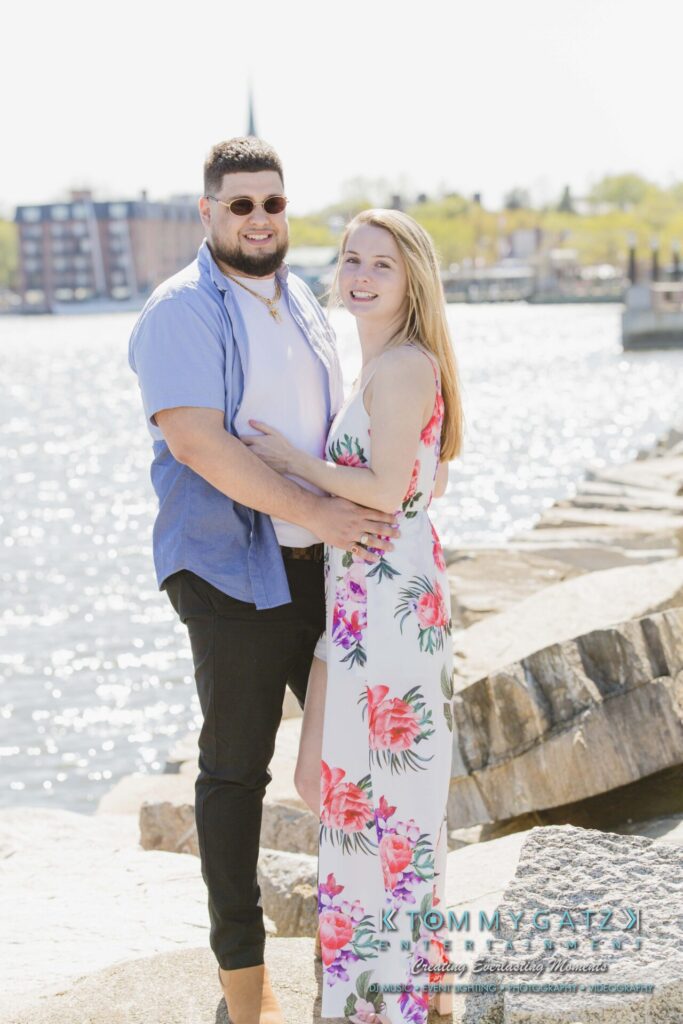 A couple stands on a rocky waterfront path, with the man in a blue shirt and the woman in a floral dress, smiling at the camera.