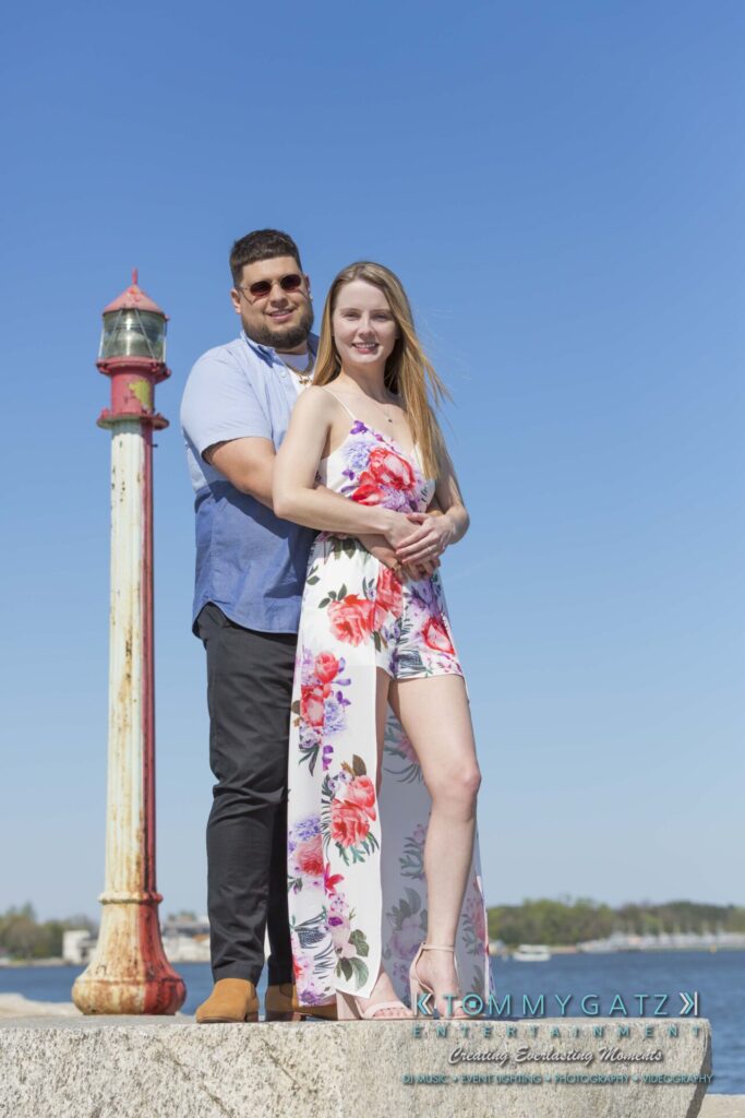 A couple stands together on a pier with a red and white post in front. The woman wears a floral dress, and the man wears a blue shirt and sunglasses. A body of water is in the background.