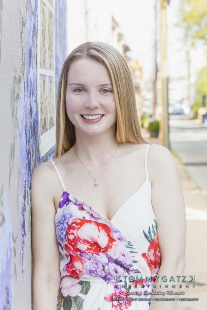 A woman in a floral dress stands next to a painted wall on a sunny day, smiling at the camera.