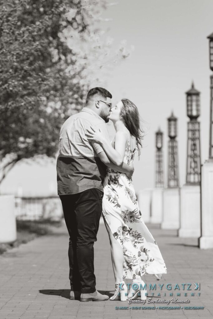 A couple embraces while standing on a paved path lined with trees and lampposts. The woman wears a floral dress, and the man wears a short-sleeved shirt.