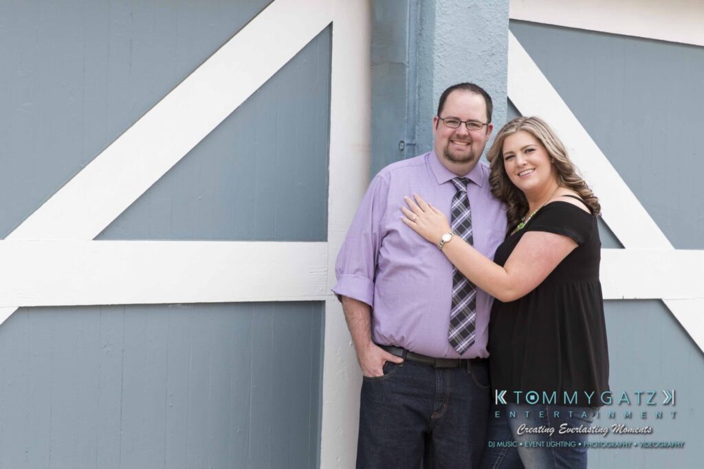 A man and woman pose together against a blue and white barn door. The woman rests her hand on the man's shoulder. Both are smiling.